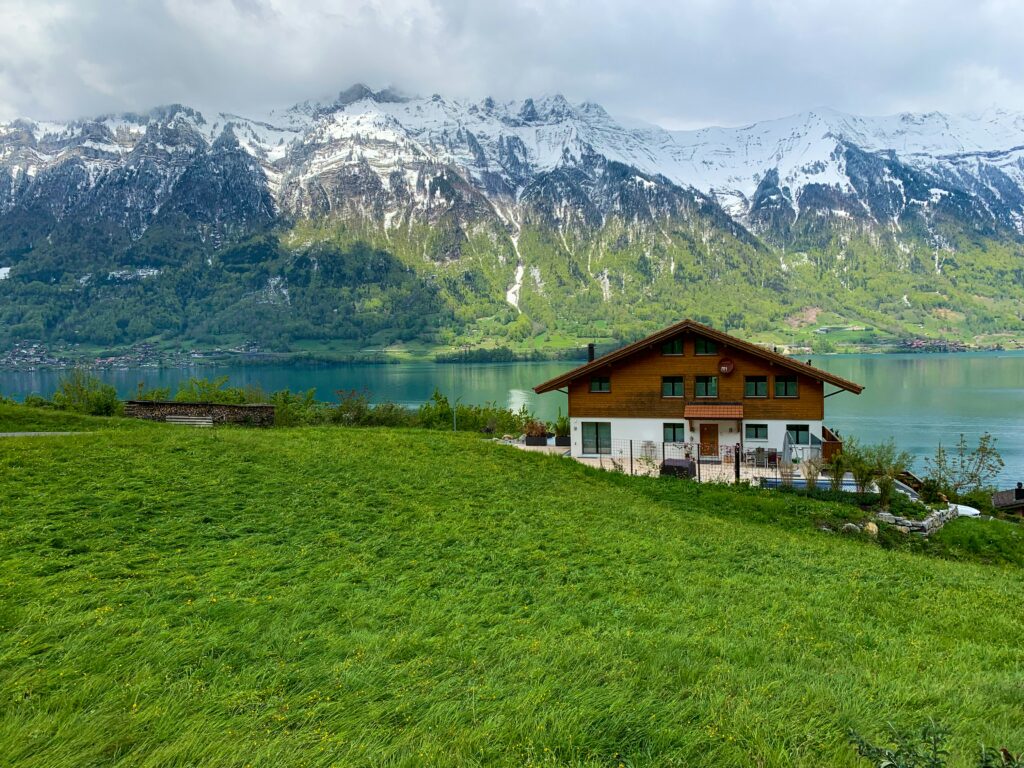 Schweizer Holzchalet auf grüner Wiese am türkisblauen Bergsee mit schneebedeckten Alpen im Hintergrund