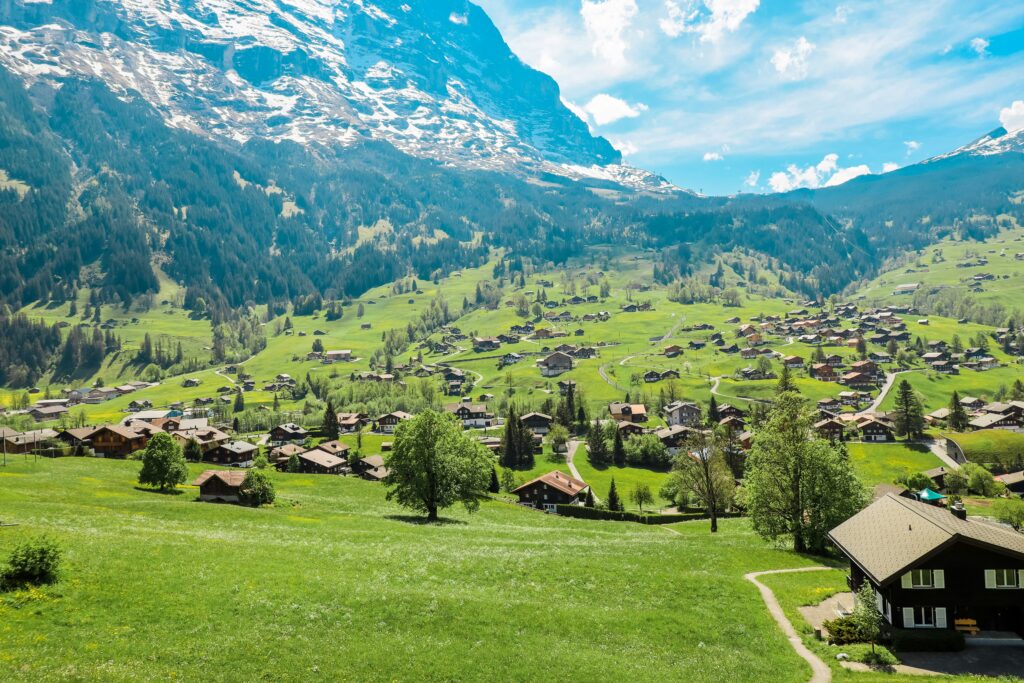 **Alt-Tag:** `Luftaufnahme des Schweizer Bergdorfs Grindelwald im Berner Oberland: grüne Almwiesen mit verstreuten Holzchalets, umgeben von bewaldeten Hängen und schneebedeckten Gipfeln der Berner Alpen unter blauem Himmel`