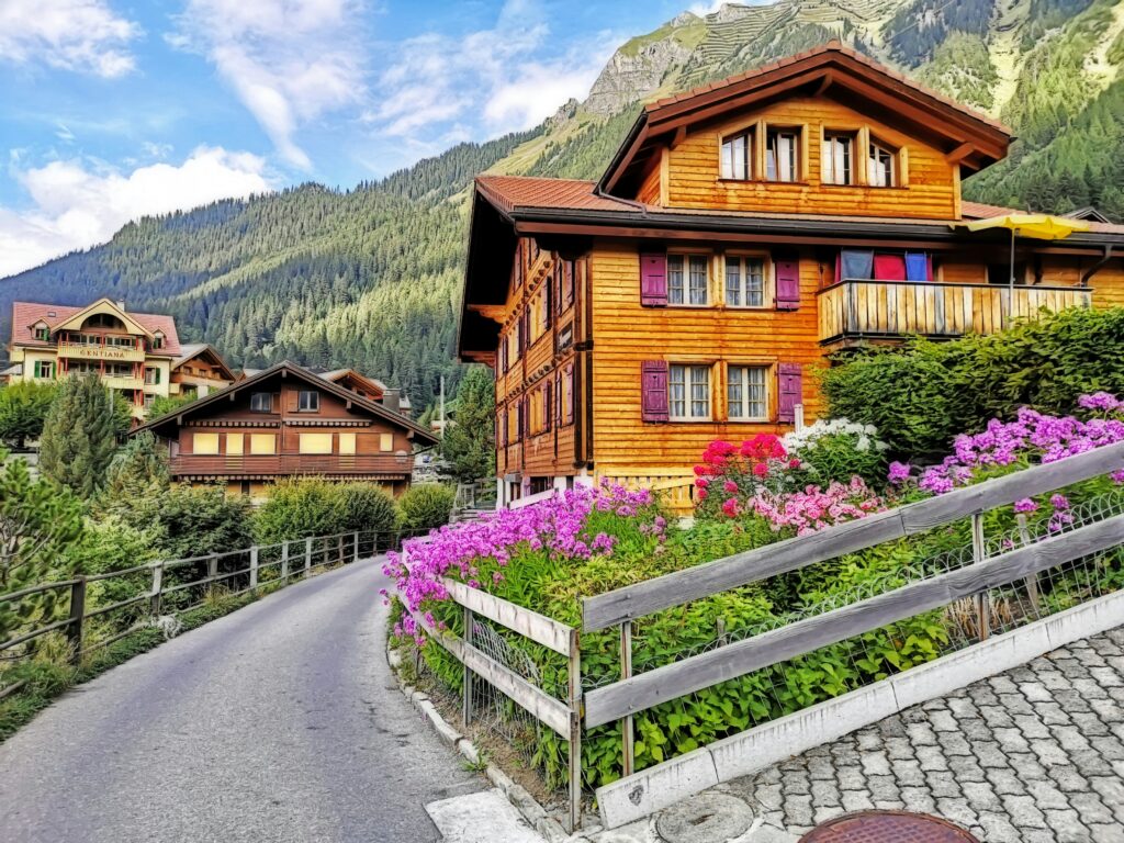 Traditionelles Schweizer Holzchalet mit lila Fensterläden und blühenden Blumen an einer Bergstraße