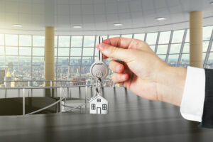 Hand übergibt Wohnungsschlüssel mit Hausanhänger vor Panoramafenster mit Stadtblick