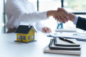 Immobiliendeal wird per Handschlag besiegelt, gelbes Hausmodell im Vordergrund