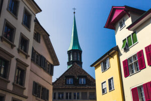 Grüner Kirchturm zwischen bunten Altstadthäusern mit roten und grünen Fensterläden vor blauem Himmel