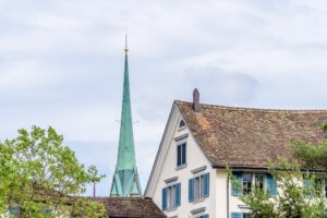 Grün patinierte Kirchturmspitze ragt hinter dem Giebel eines historischen Hauses mit rotbraunem Ziegeldach und blauen Fensterläden hervor