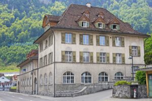 Historisches mehrstöckiges Gebäude mit grünen Fensterläden, Walmdach und Gauben vor bewaldetem Hang