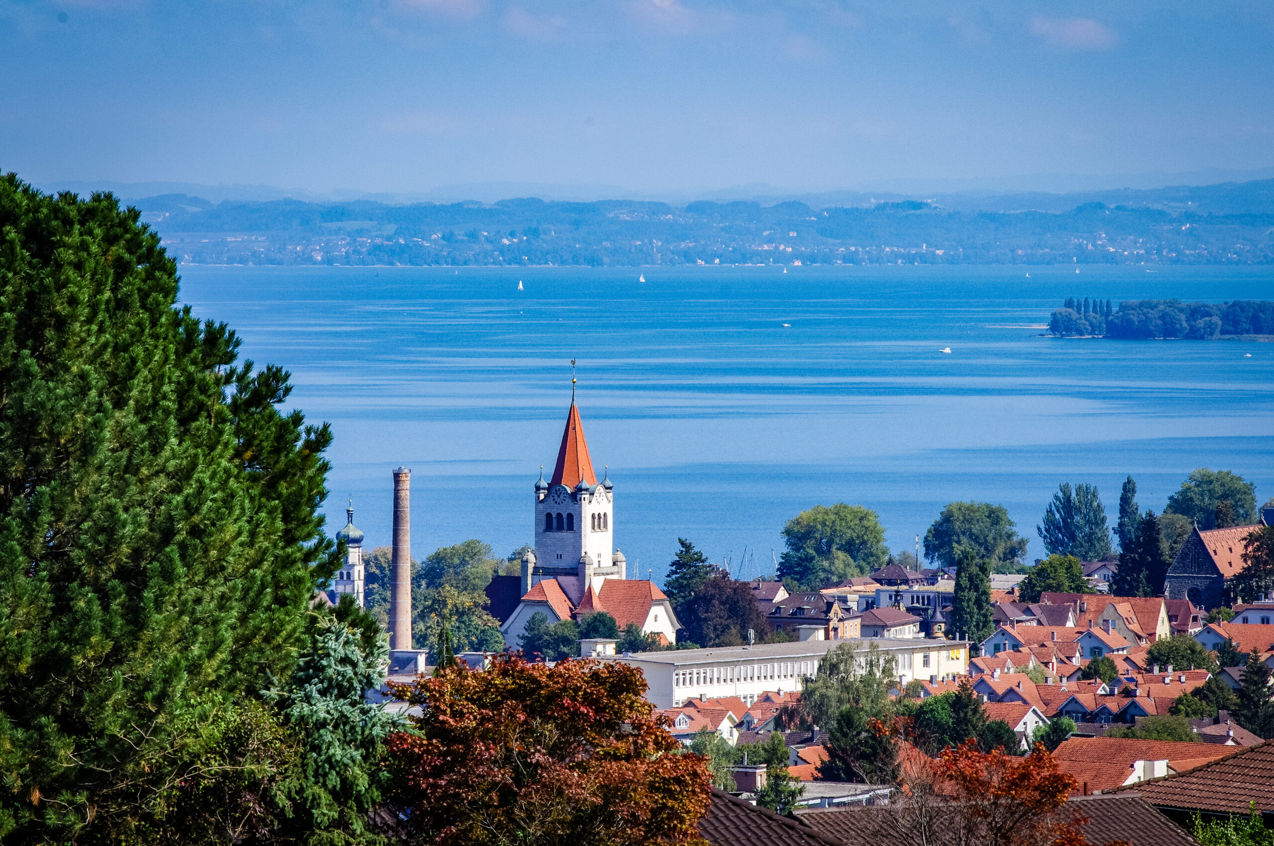 Blick über Dächer und einen Kirchturm mit rotem Spitzdach auf den Bodensee mit Segelbooten und Hügeln am Horizont