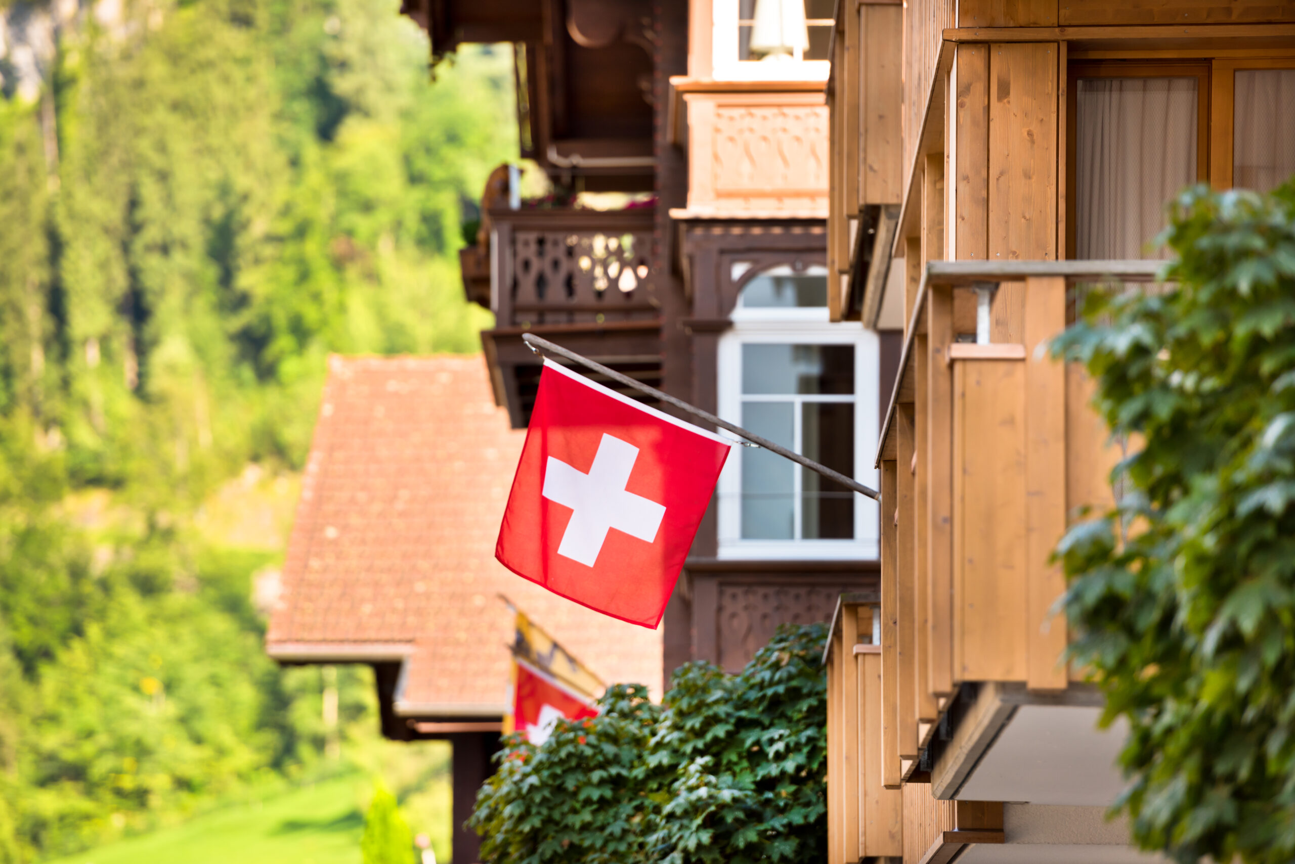 chweizer Flagge weht an einem traditionellen Holzchalet mit Balkon, im Hintergrund bewaldete Berghänge